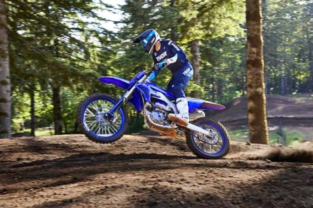 A motocross rider in a blue THOR kit performs a jump on a Yamaha YZ450F dirt bike on a forested trail.
