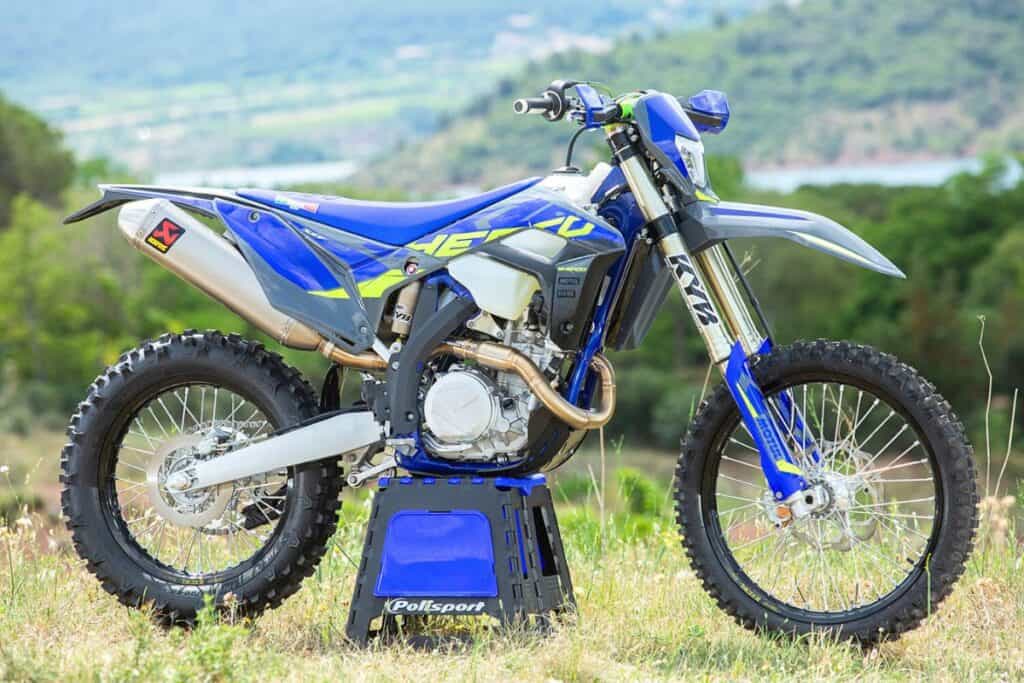 A Sherco 450 SEF dirt bike is perched on a blue Polisport stand in a grassy field with a scenic hilly backdrop.