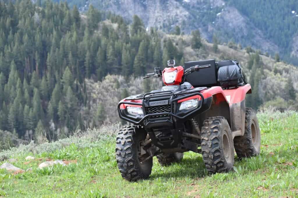 A red Honda ATV equipped with hunting accessories is parked on a grassy clearing with a dense forest backdrop.