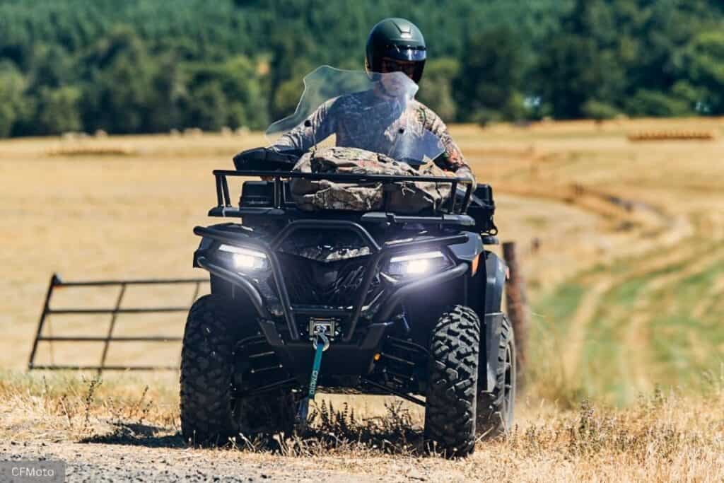 A person wearing a helmet and camo gear is riding a CFMoto CForce 600 hunting ATV across a dry, grassy field.