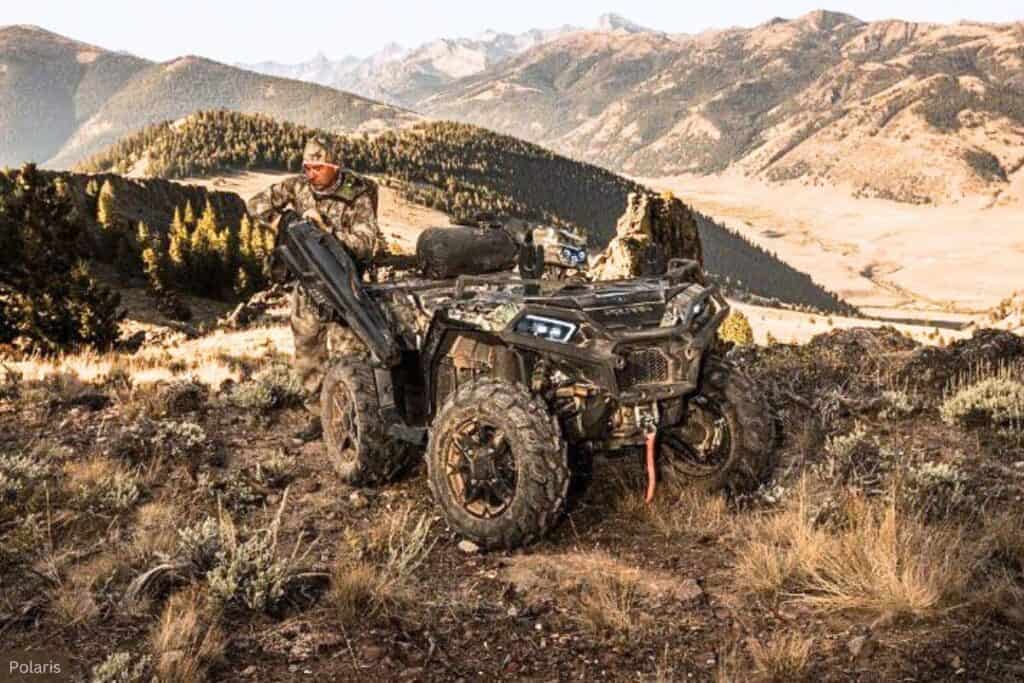 A hunter in camouflage is standing next to a Polaris Sportsman XP 1000 Hunt Edition on a mountain overlook with a panoramic view of forested hills.
