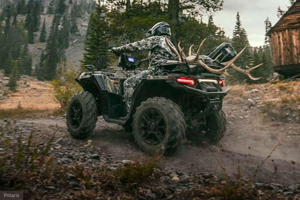 A rider dressed in camouflage gear is hunting with their Polaris Sportsman 570 Hunt Edition ATV, carrying antlers on the back, on a rugged mountain trail.